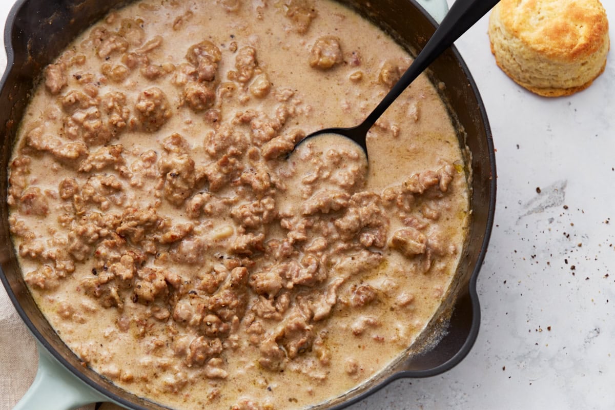 Close-up of a spoon dipping into thick, creamy gluten-free sausage gravy in a skillet, with a biscuit nearby.