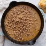 Overhead view of a cast iron skillet filled with thick, creamy gluten-free sausage gravy, with golden biscuits on the side.