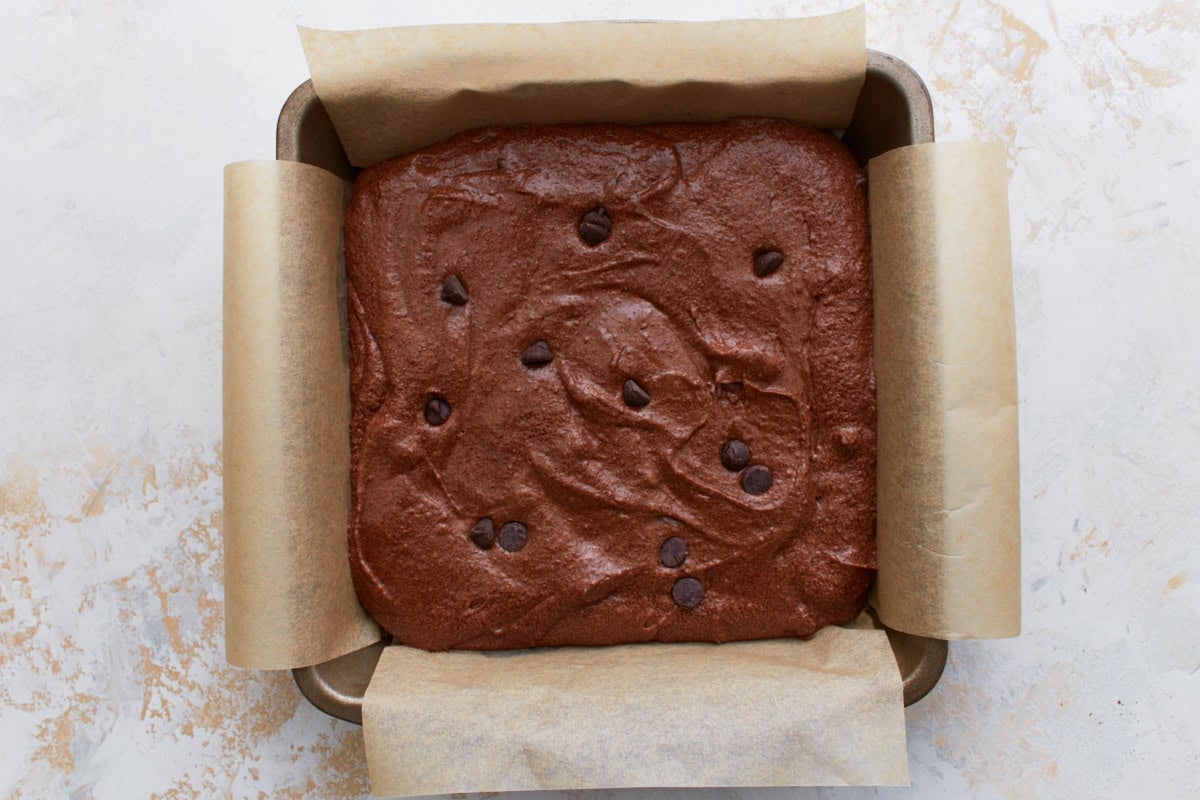 Raw keto brownie batter spread evenly in a parchment-lined square pan, topped with extra chocolate chips.