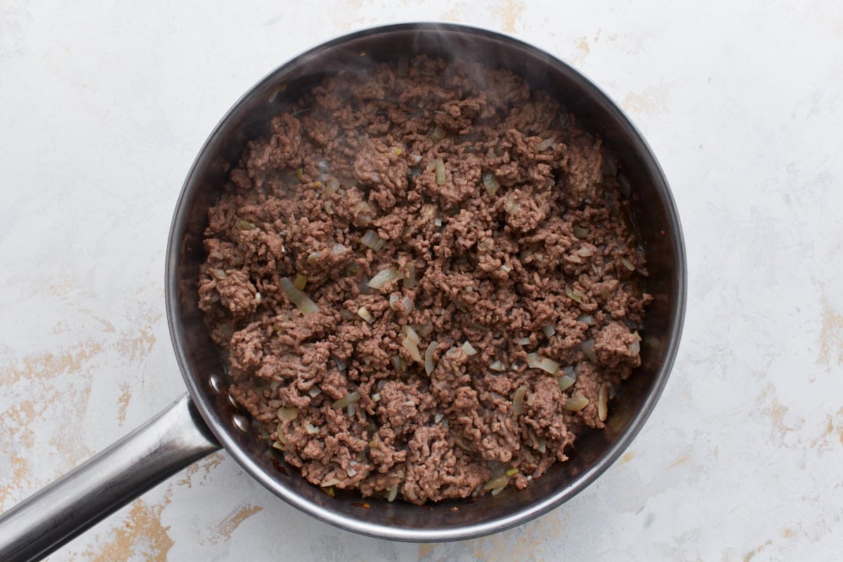 Ground beef cooking with softened onions and garlic in a skillet, starting to brown and break apart.