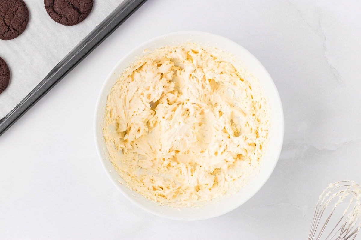 A white bowl filled with fluffy vanilla marshmallow cream filling, ready to be piped onto cookies. A baking tray with chocolate cookies and a whisk are partially visible nearby.