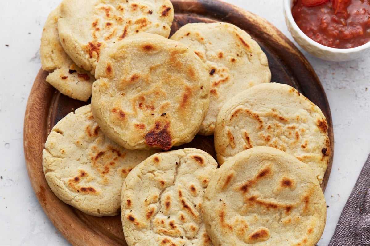 A wooden platter filled with cooked pupusas, golden and slightly crisp, with a bowl of salsa off to the side.