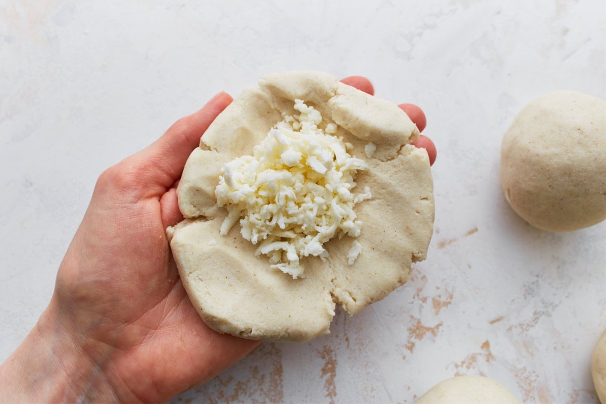 Alt text: A close-up of a hand holding flattened masa dough topped with a pile of shredded white cheese.