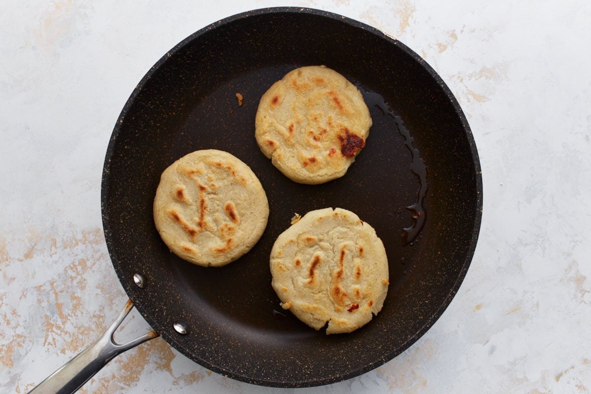 Three pupusas cooking in a skillet, lightly browned and developing charred spots.