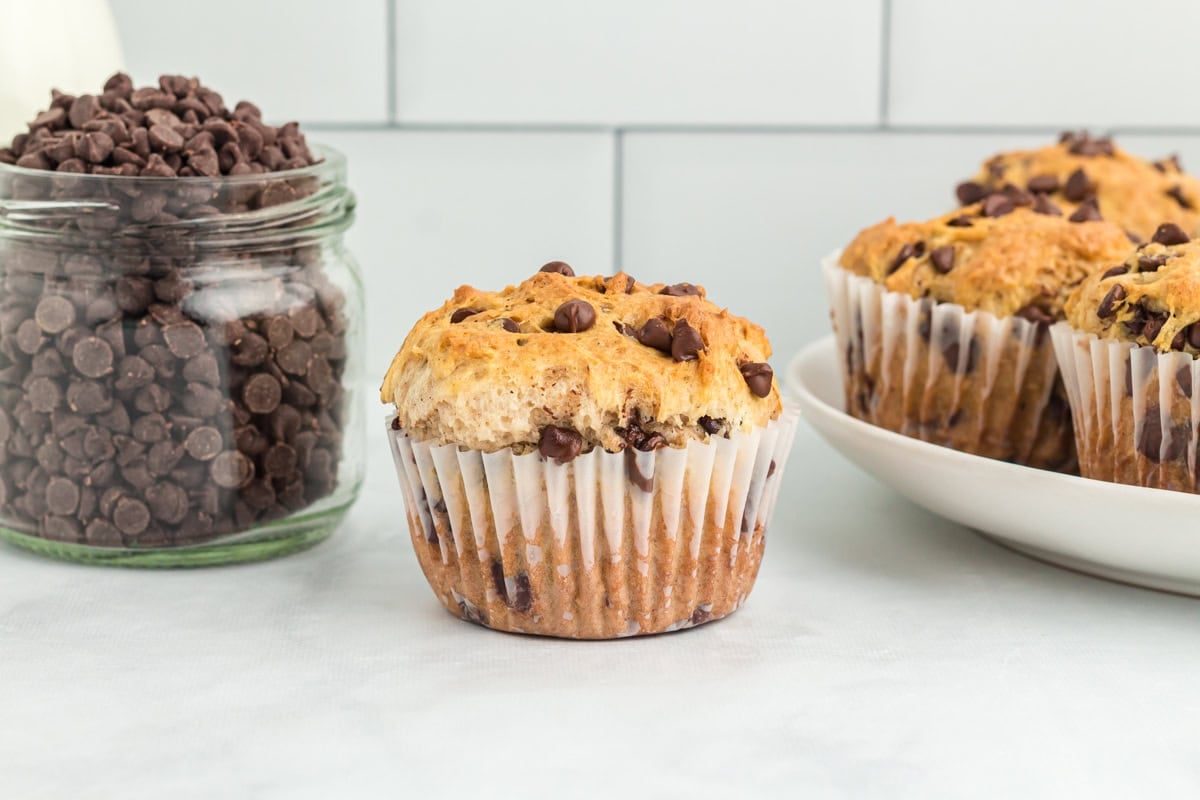 A gluten-free chocolate chip muffin in a white paper liner placed in front of a glass jar filled with chocolate chips, with more muffins on a plate in the background.