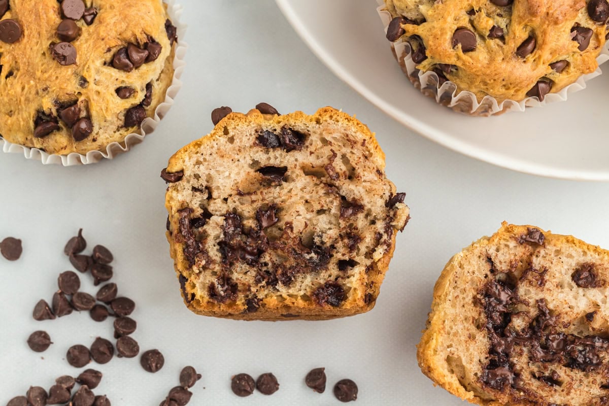 Overhead view of halved gluten-free chocolate chip muffins showing a soft, fluffy interior filled with melted chocolate chips, with scattered chocolate chips on a white surface nearby.