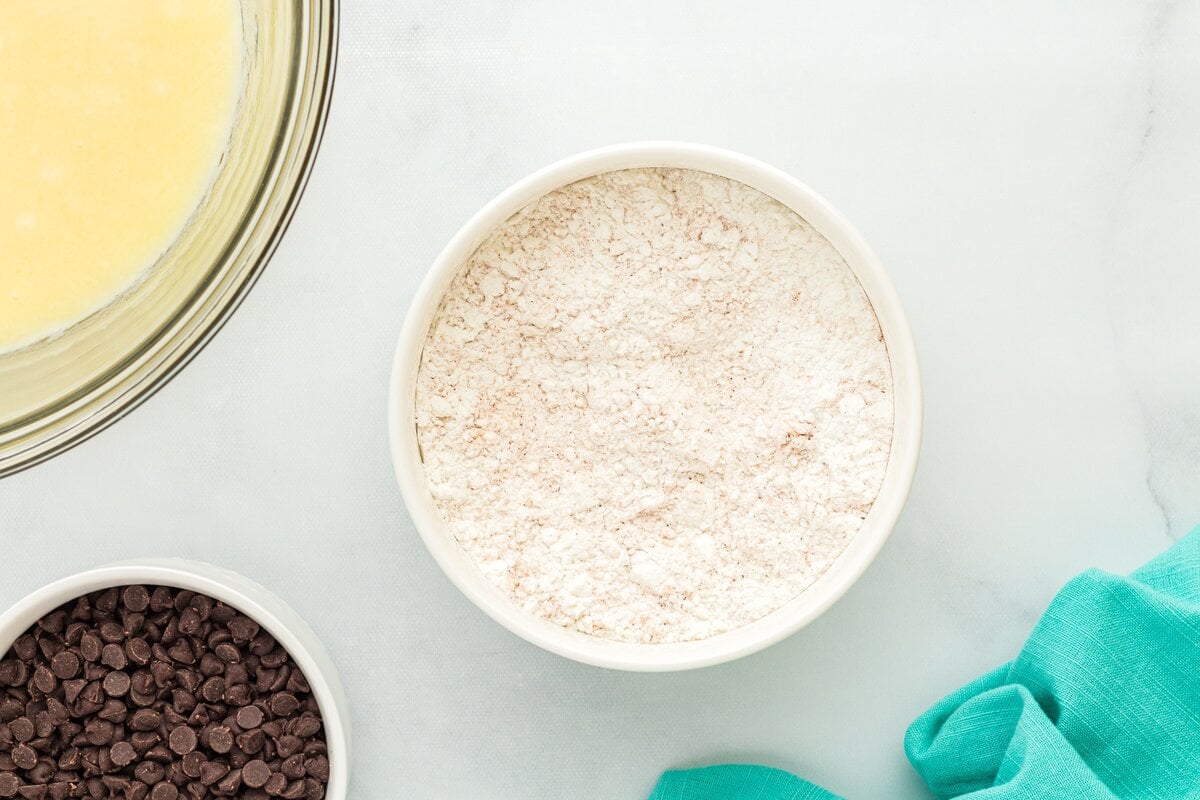 A bowl of gluten-free flour next to a bowl of chocolate chips and a glass bowl of wet muffin ingredients, set on a white surface with a teal towel.