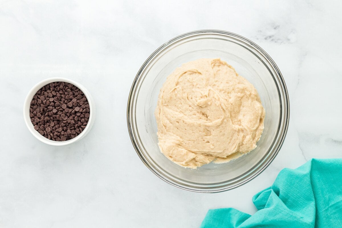 A bowl of muffin batter before the chocolate chips have been added, sitting beside a bowl of mini chocolate chips.