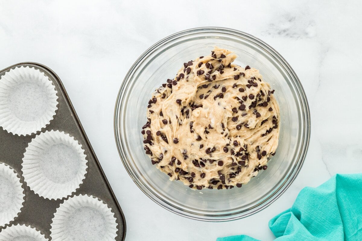A bowl of muffin batter fully mixed with mini chocolate chips, next to a muffin tin lined with paper wrappers.