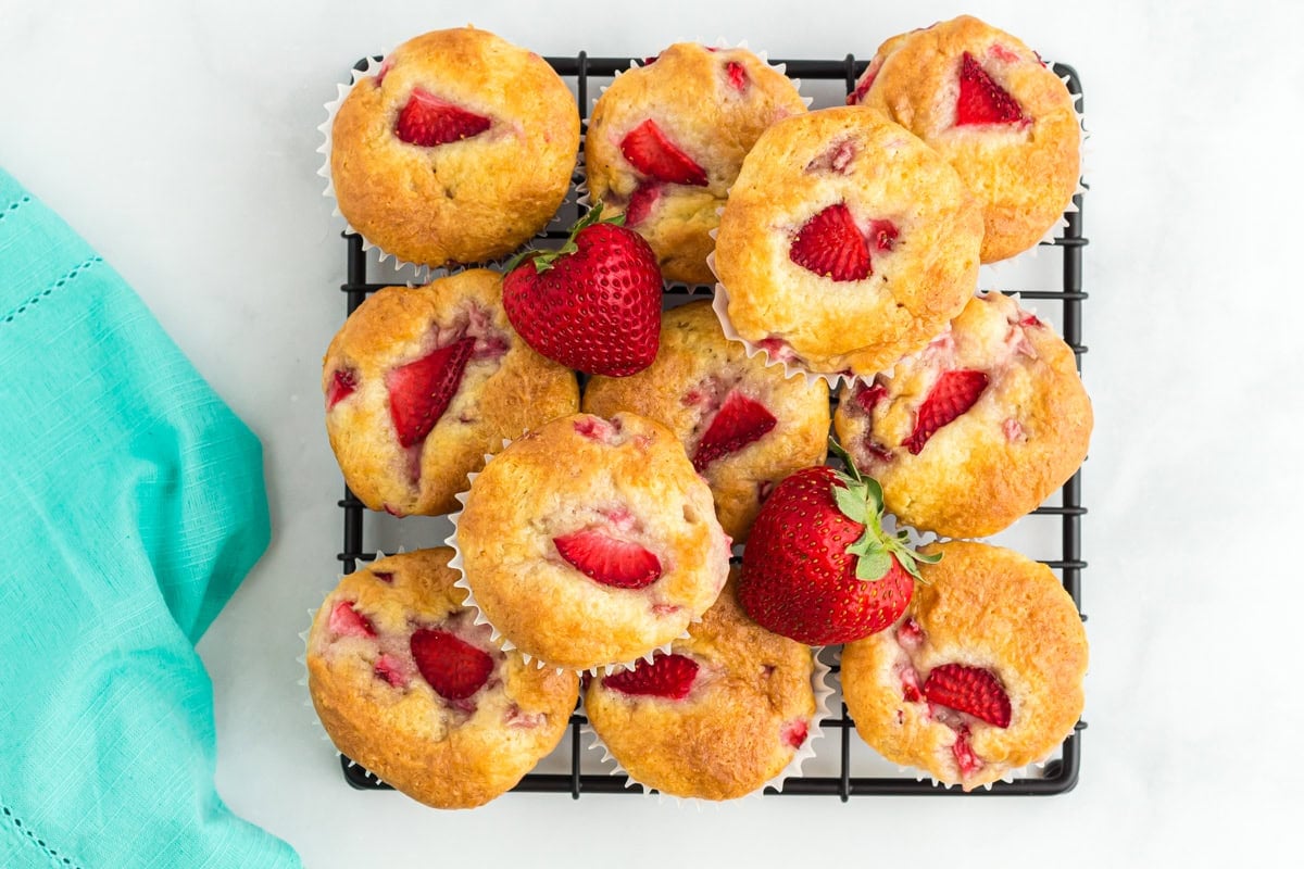 Overhead view of a pile of golden gluten-free strawberry muffins on a black cooling rack, garnished with fresh whole strawberries.