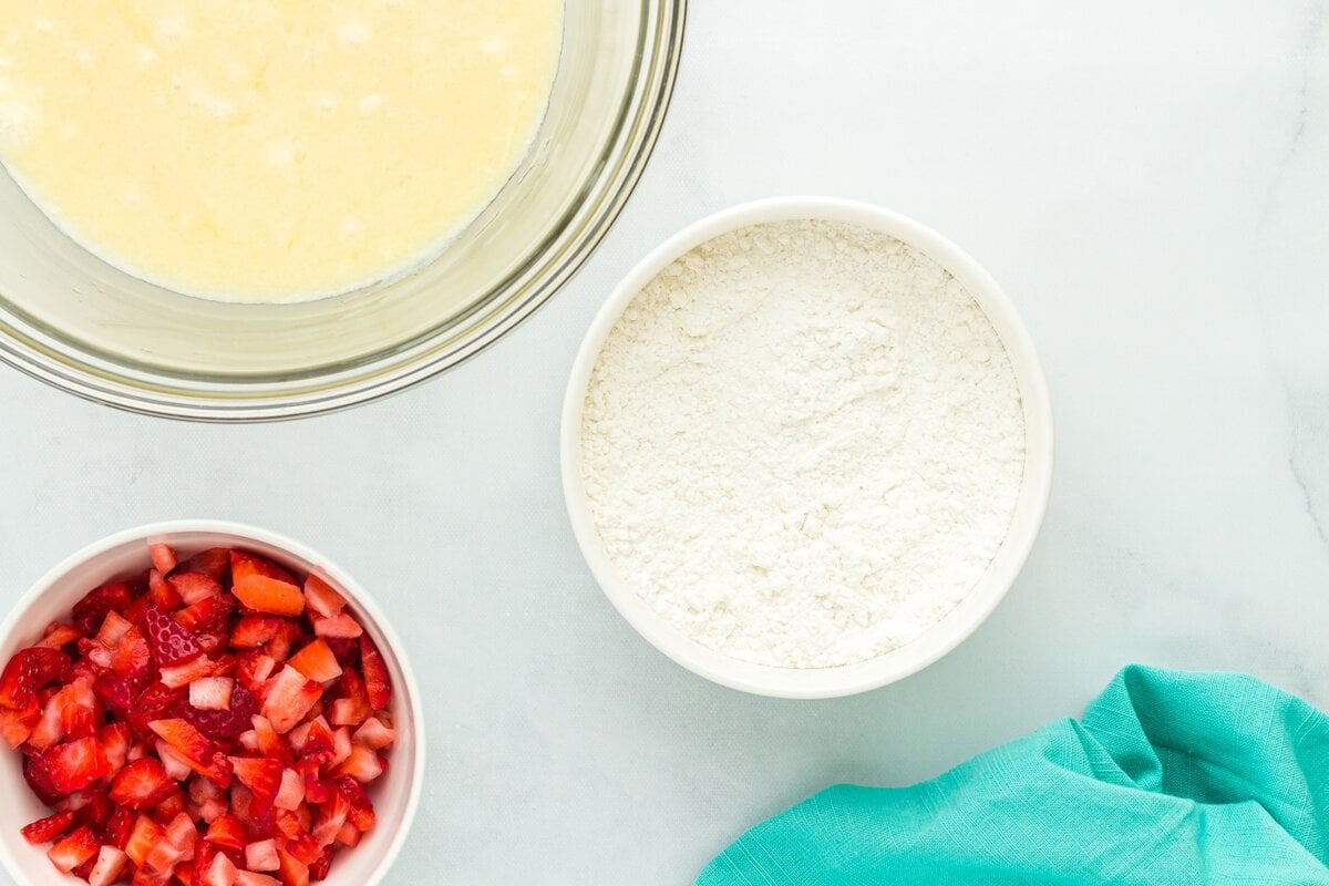 Three bowls on a light background: one with chopped fresh strawberries, one with gluten-free flour, and one with a whisked wet mixture.