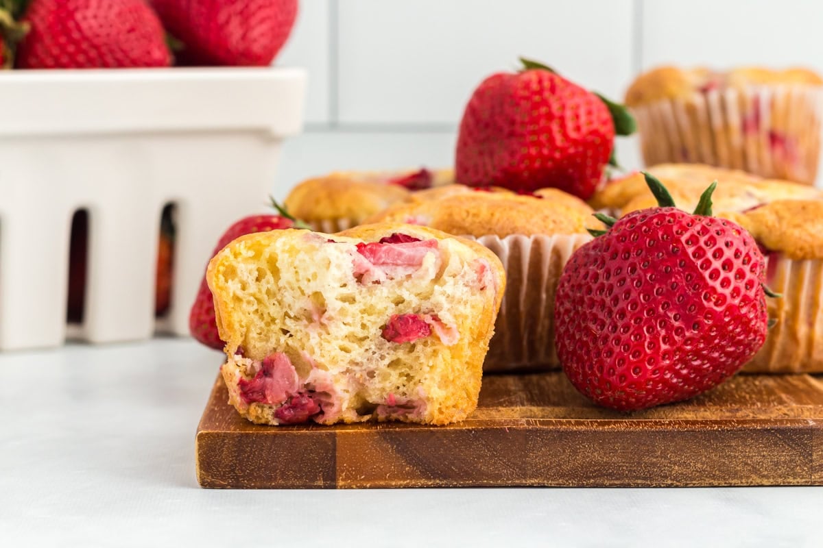 A close-up of a gluten-free strawberry muffin with a bite taken out, showing the fluffy interior and chunks of fresh strawberries, surrounded by whole strawberries and additional muffins in the background
