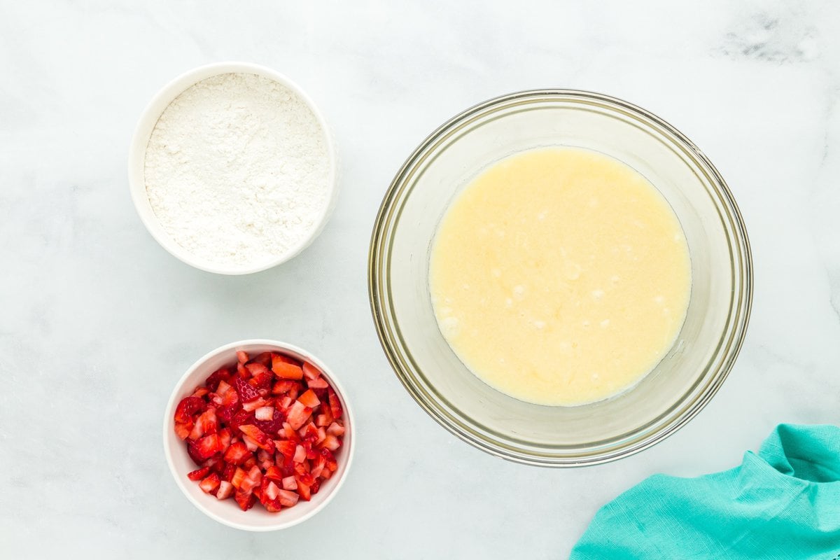 Top-down view of three bowls containing chopped strawberries, gluten-free flour, and wet muffin batter, set on a white surface