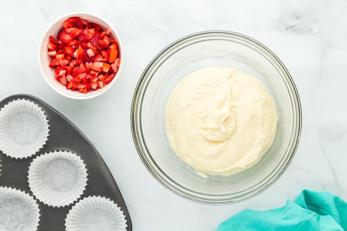 A bowl of thick muffin batter next to a bowl of chopped strawberries and a lined muffin pan, ready for assembly.