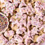 A plate of gluten-free animal crackers, some dipped in pink candy coating and sprinkled with rainbow nonpareils, with a small dish of extra sprinkles on the side.