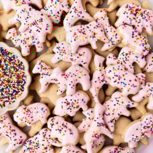 A plate of gluten-free animal crackers, some dipped in pink candy coating and sprinkled with rainbow nonpareils, with a small dish of extra sprinkles on the side.