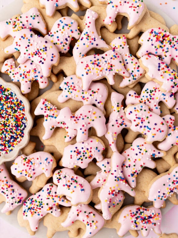 A plate of gluten-free animal crackers, some dipped in pink candy coating and sprinkled with rainbow nonpareils, with a small dish of extra sprinkles on the side.
