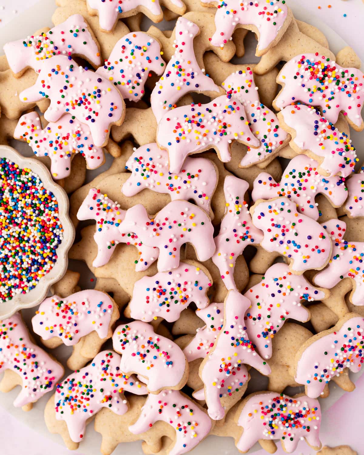 A plate of gluten-free animal crackers, some dipped in pink candy coating and sprinkled with rainbow nonpareils, with a small dish of extra sprinkles on the side.