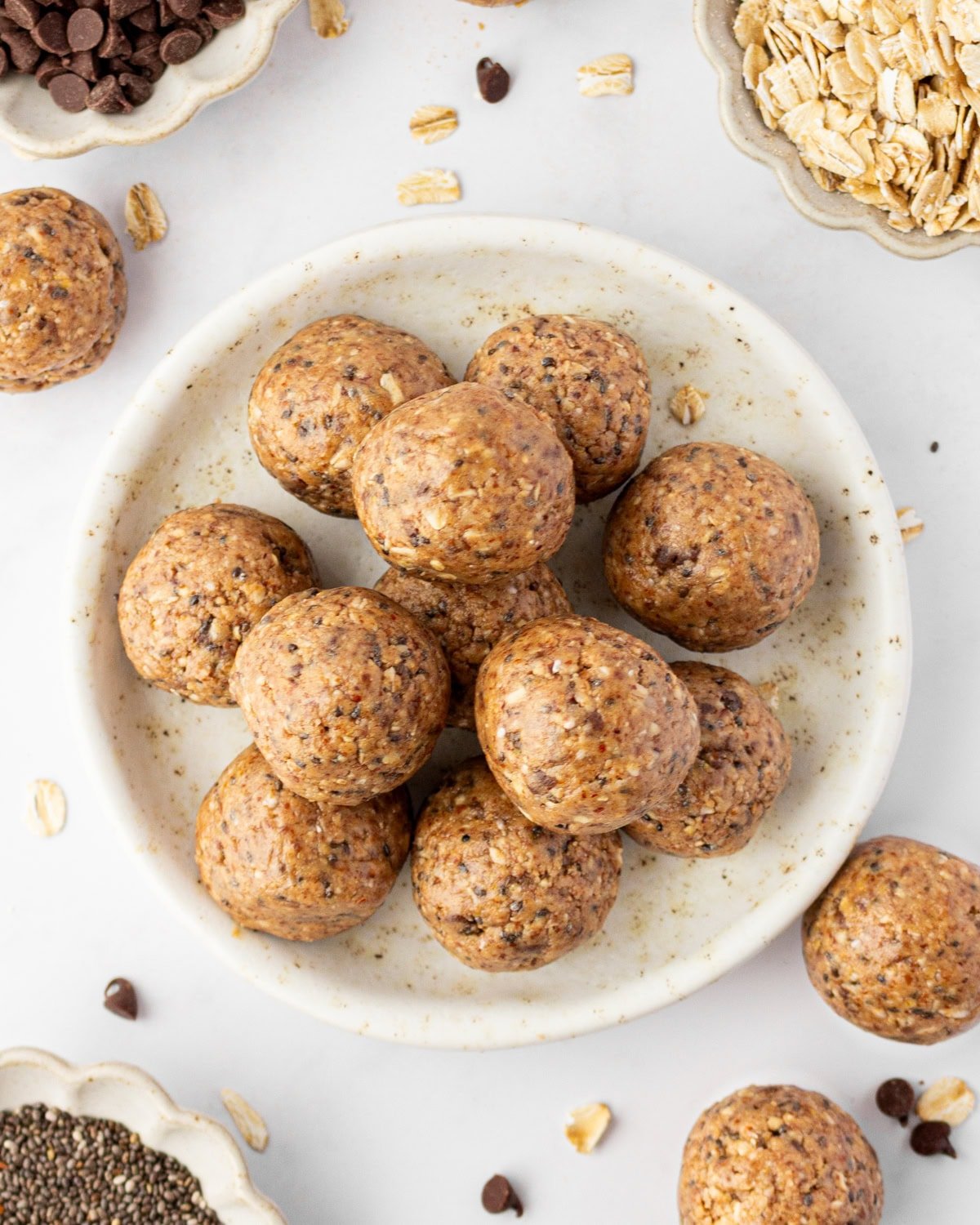A plate of gluten free protein balls stacked in the center, surrounded by small bowls of chocolate chips, rolled oats, and chia seeds.