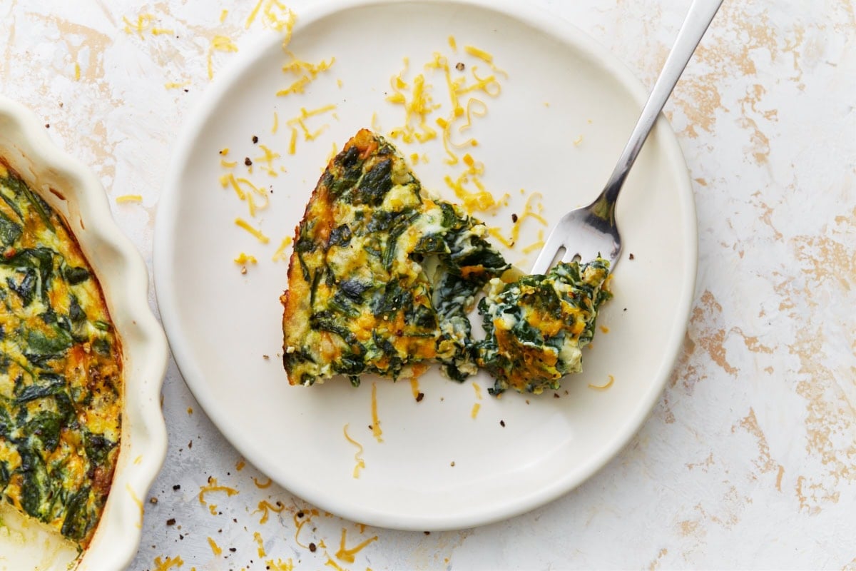 Slice of crustless spinach quiche on a white plate with a fork.