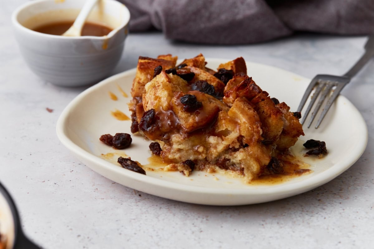 A serving of gluten-free bread pudding with raisins on a white plate, drizzled with caramel sauce, with a fork beside it.