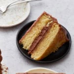A slice of moist, two-layer gluten-free yellow cake with chocolate buttercream frosting on a small black plate, with an empty plate and forks in the background.