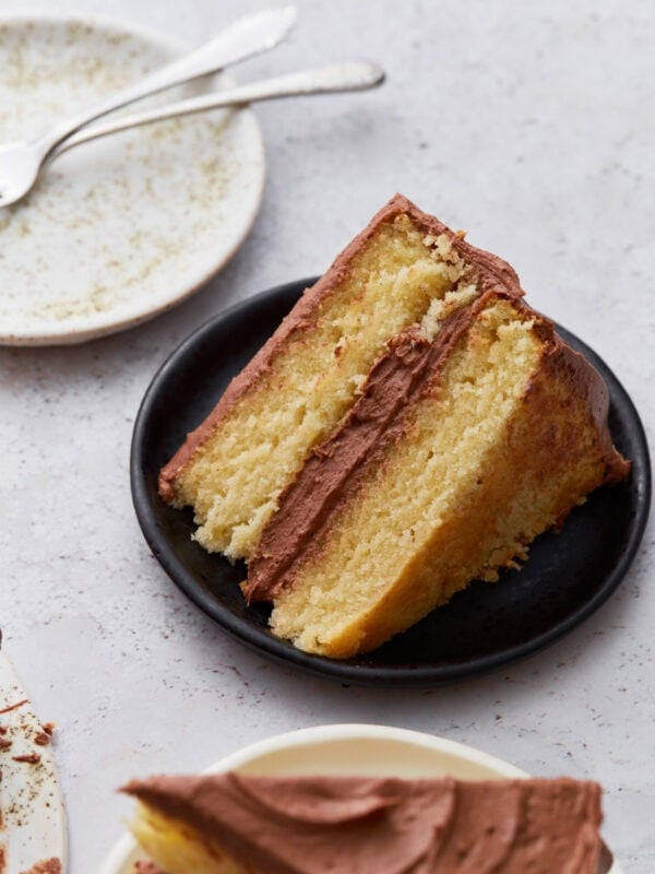 A slice of moist, two-layer gluten-free yellow cake with chocolate buttercream frosting on a small black plate, with an empty plate and forks in the background.