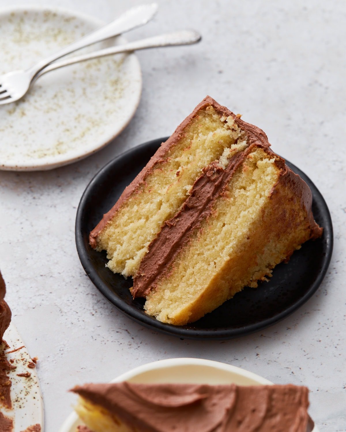 A slice of moist, two-layer gluten-free yellow cake with chocolate buttercream frosting on a small black plate, with an empty plate and forks in the background.