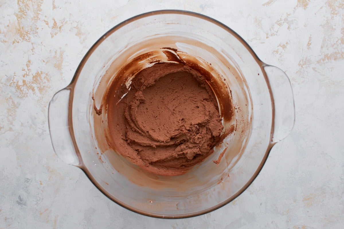 Cocoa powder and other frosting ingredients partially mixed into butter, forming a thick chocolate base in a glass bowl.