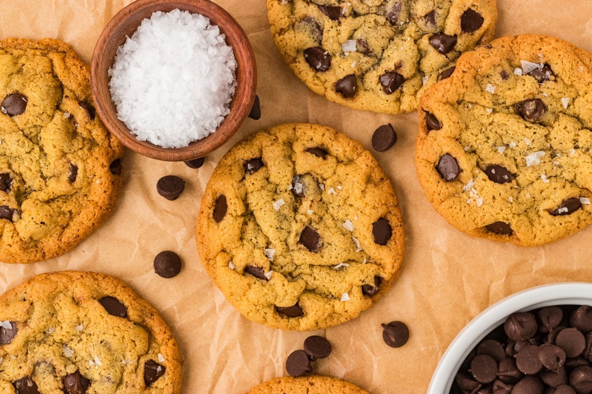 Overhead view of chickpea chocolate chip cookies sprinkled with flaky salt, surrounded by chocolate chips and a small bowl of salt.