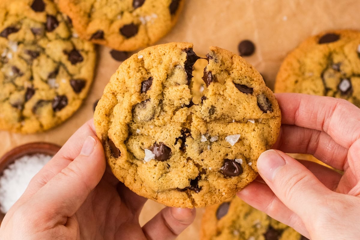 Hands breaking apart a chickpea chocolate chip cookie topped with flaky salt, with melted chocolate visible inside.