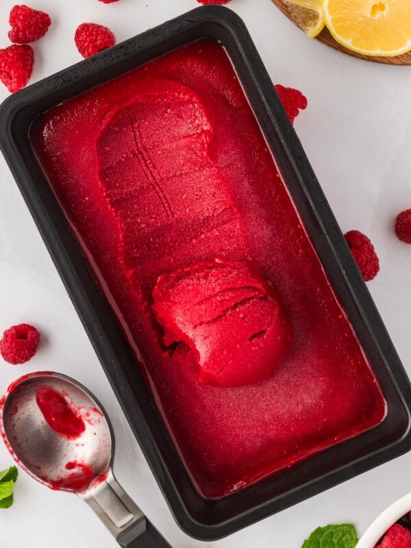 A black loaf pan filled with bright red raspberry sorbet, with one scoop removed and a metal ice cream scoop resting beside fresh raspberries and lemon slices.