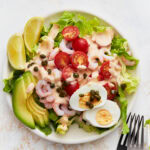 A plate of Shrimp Louie salad with romaine, bay shrimp, cherry tomatoes, avocado slices, hard-boiled egg, capers, and Louie dressing, served with lemon wedges.
