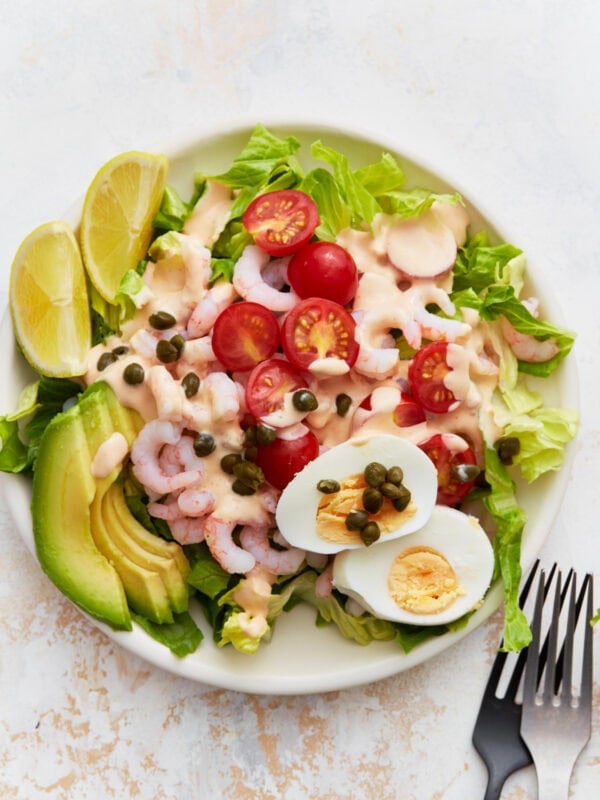 A plate of Shrimp Louie salad with romaine, bay shrimp, cherry tomatoes, avocado slices, hard-boiled egg, capers, and Louie dressing, served with lemon wedges.