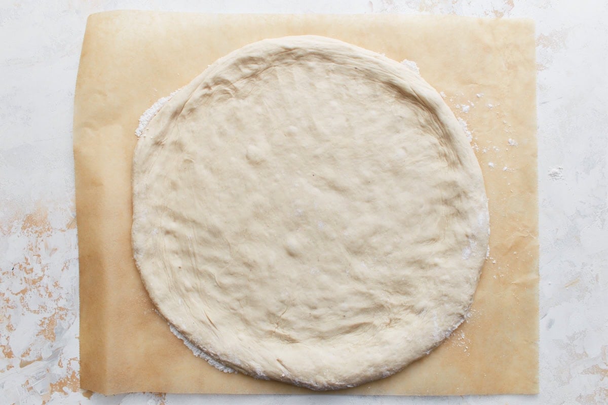 The pizza dough flattened into a 12-inch circle on parchment paper, ready for sauce and toppings.