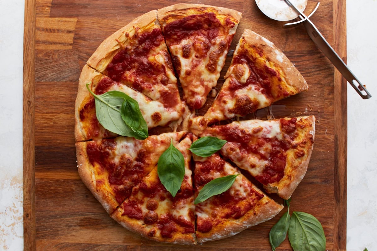 The baked vegan pizza cut into slices and topped with fresh basil, a pizza cutter nearby on a wooden board.