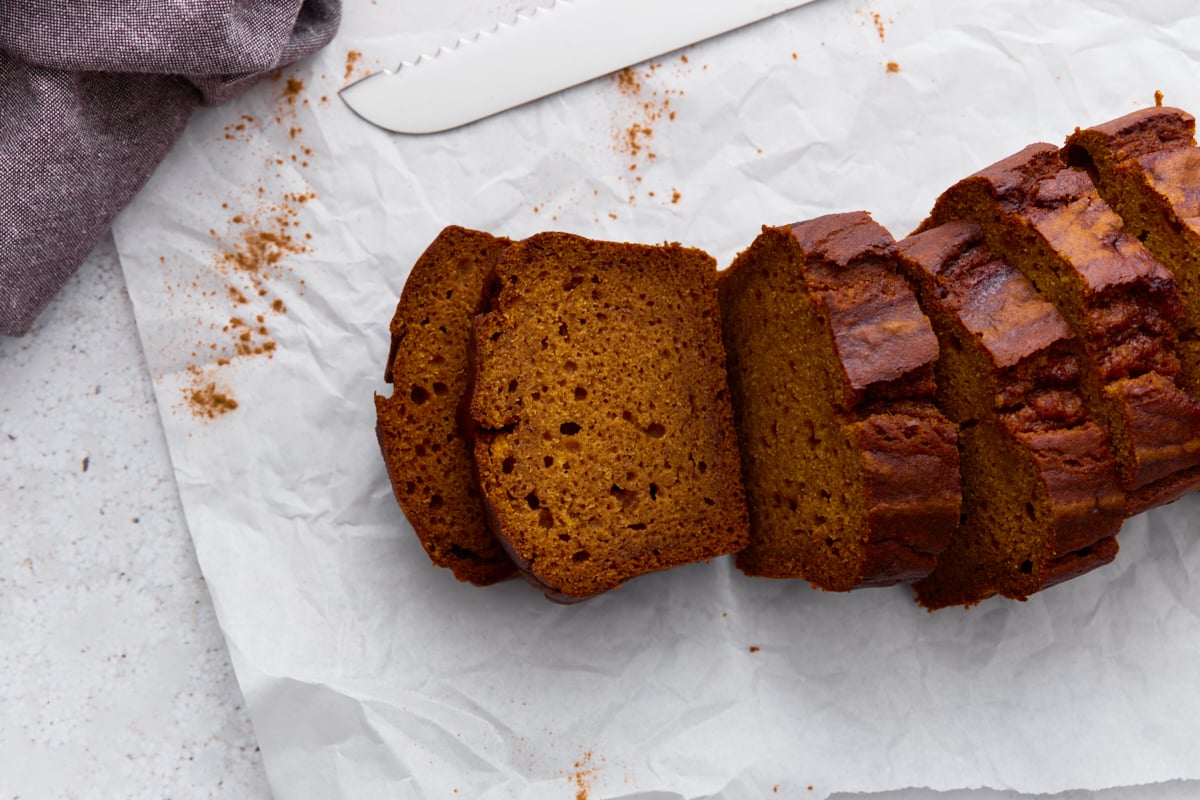 Pumpkin bread sliced on parchment paper with a serrated knife nearby, crumbs scattered.