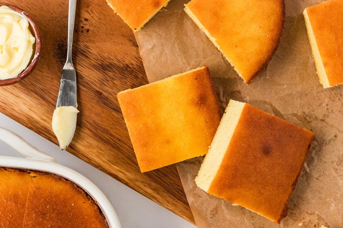 Cut squares of baked keto cornbread on a wooden board with butter on the side, ready to serve.