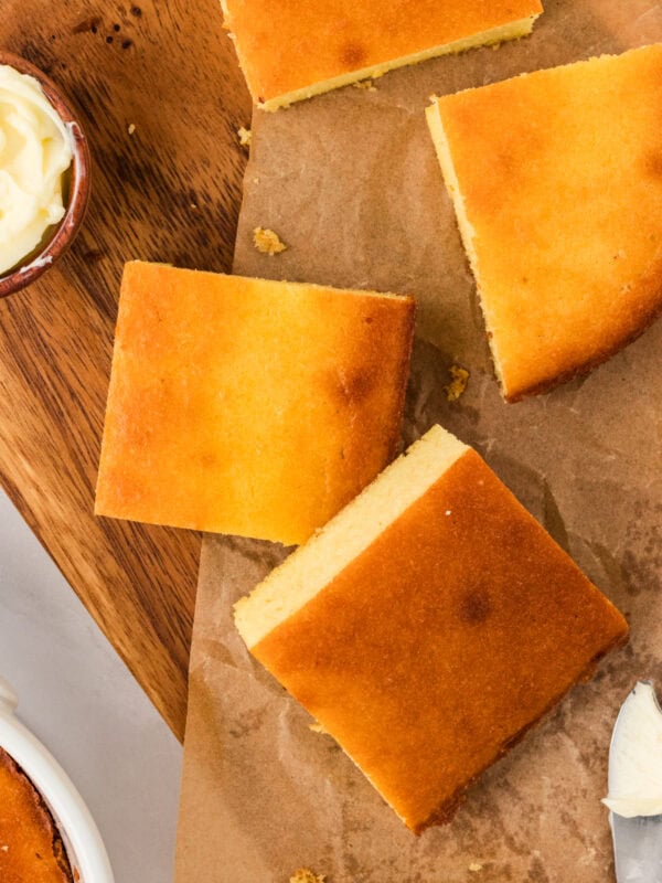 Overhead view of keto cornbread squares on parchment paper, golden brown with a small dish of butter and a butter knife nearby.