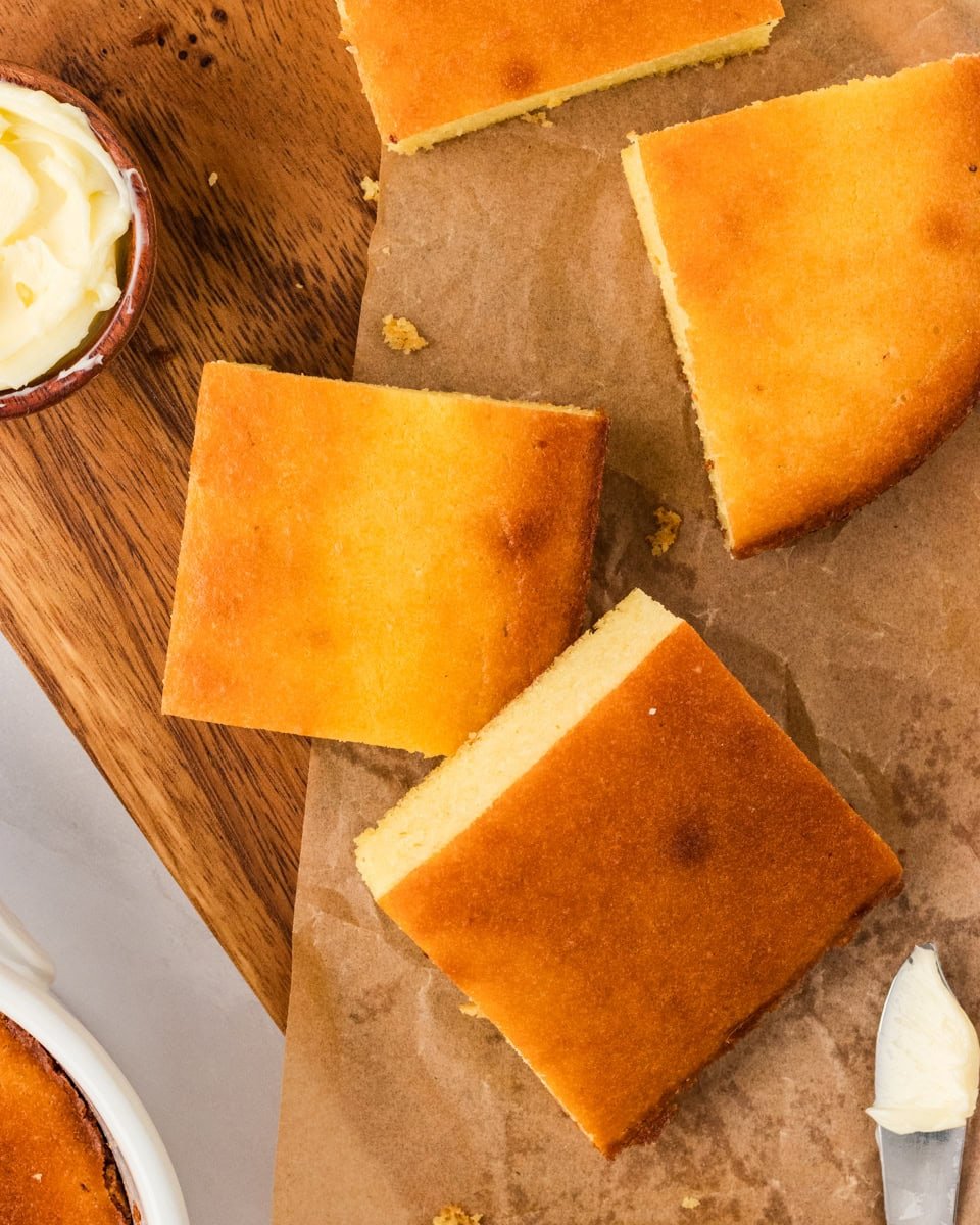 Overhead view of keto cornbread squares on parchment paper, golden brown with a small dish of butter and a butter knife nearby.