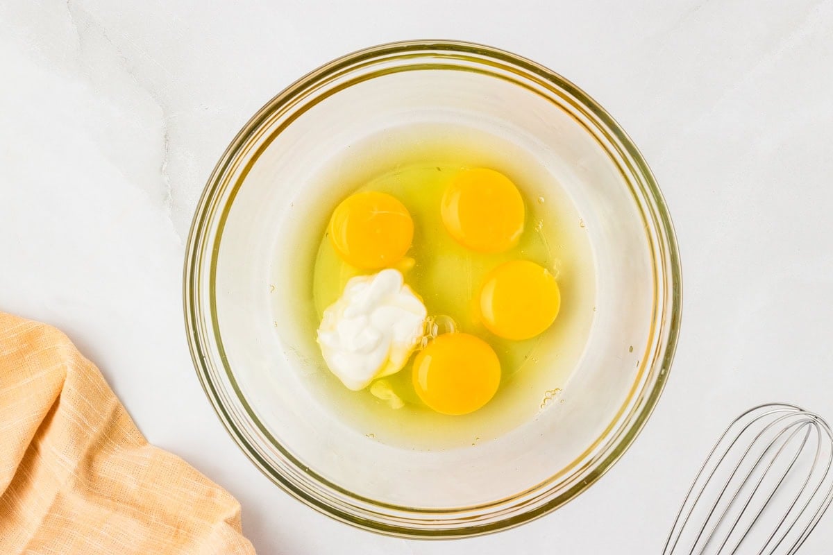 Mixing bowl with four cracked eggs and sour cream before whisking, sitting on a white counter with a whisk to the side.