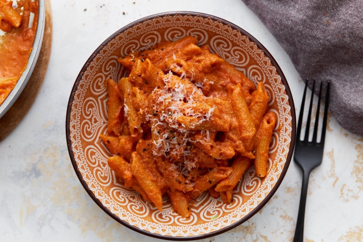 A serving of gluten-free penne pasta coated in creamy vegan vodka sauce, topped with grated vegan parmesan and cracked black pepper in a patterned bowl.