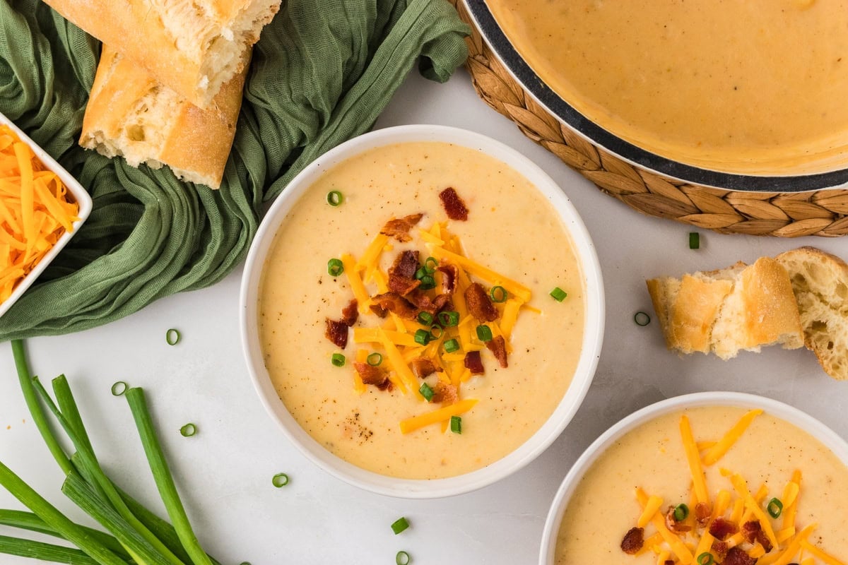 Two bowls of creamy gluten-free potato soup topped with shredded cheddar cheese, crispy bacon pieces, and chopped green onions, surrounded by a green napkin, fresh scallions, and slices of crusty bread for serving.