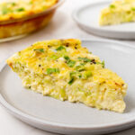 A slice of crustless zucchini quiche on a gray plate, topped with green onions, with the full quiche visible in the background.