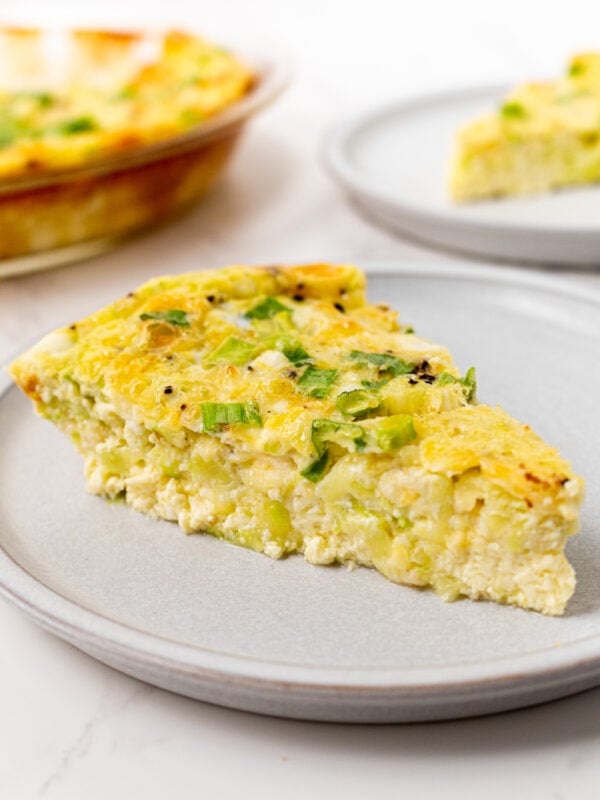 A slice of crustless zucchini quiche on a gray plate, topped with green onions, with the full quiche visible in the background.