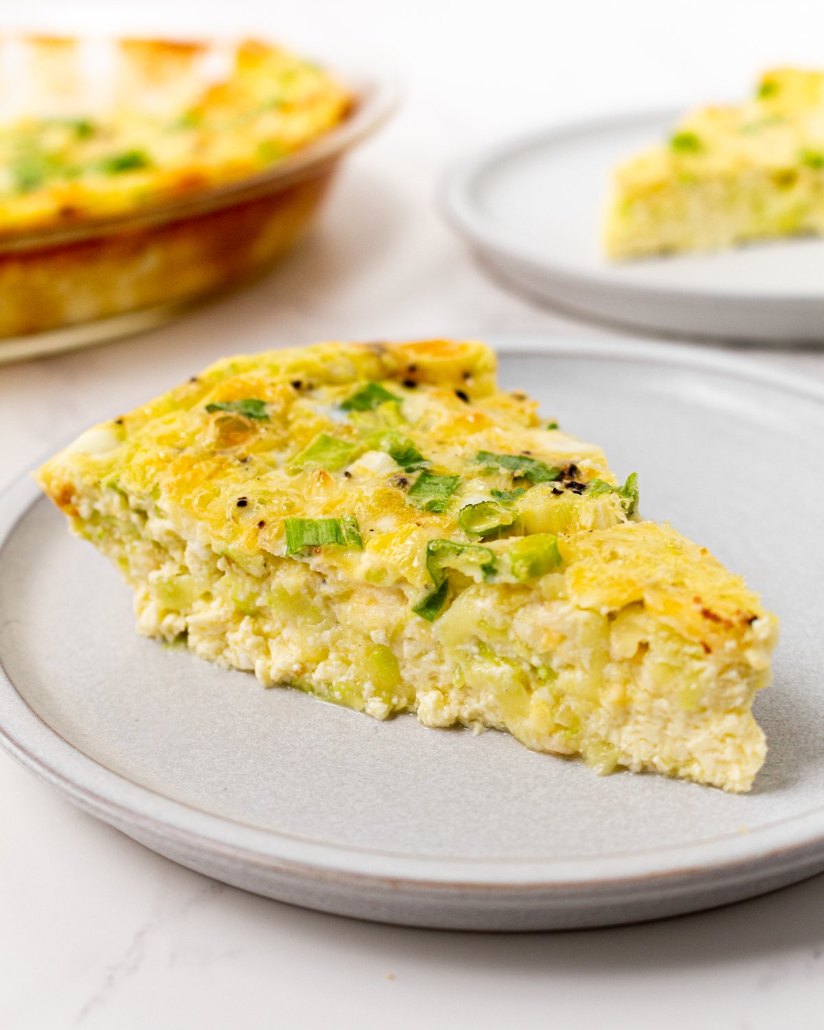 A slice of crustless zucchini quiche on a gray plate, topped with green onions, with the full quiche visible in the background.