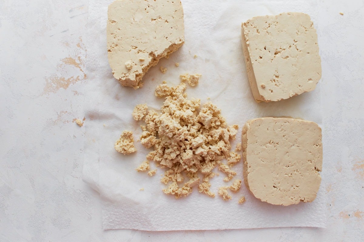 Blocks of firm tofu on a white paper towel, with some pieces crumbled into small, cottage cheese–like curds.