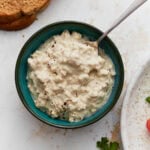 A small teal bowl filled with creamy dairy-free cottage cheese sprinkled with black pepper, with a slice of toast beside it.