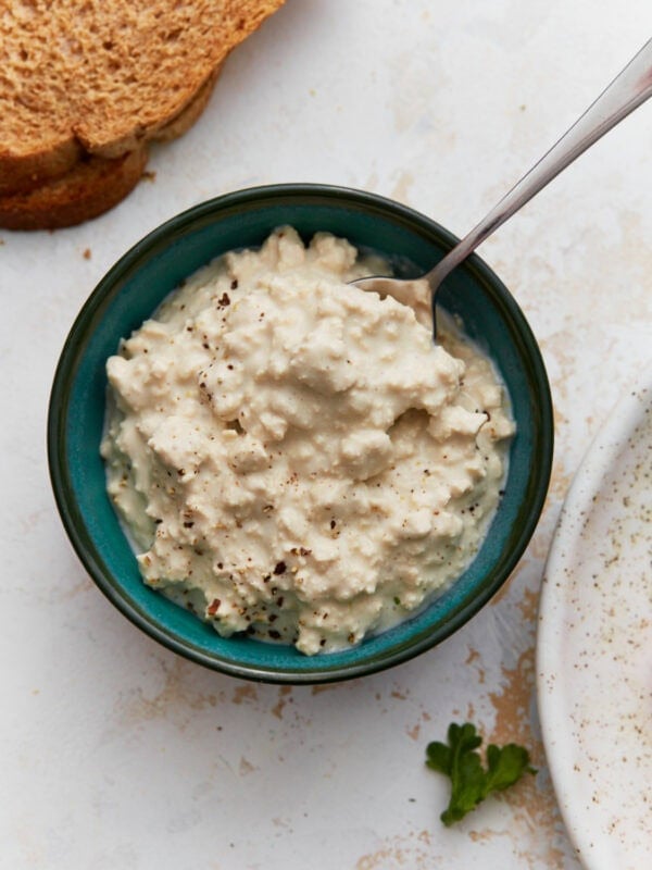 A small teal bowl filled with creamy dairy-free cottage cheese sprinkled with black pepper, with a slice of toast beside it.