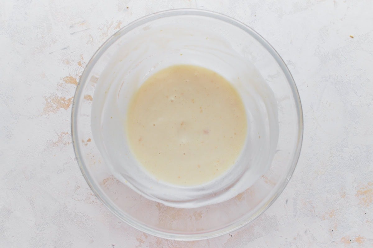 A glass mixing bowl containing a smooth, pale yellow yogurt and lemon mixture for the dairy-free cottage cheese base.